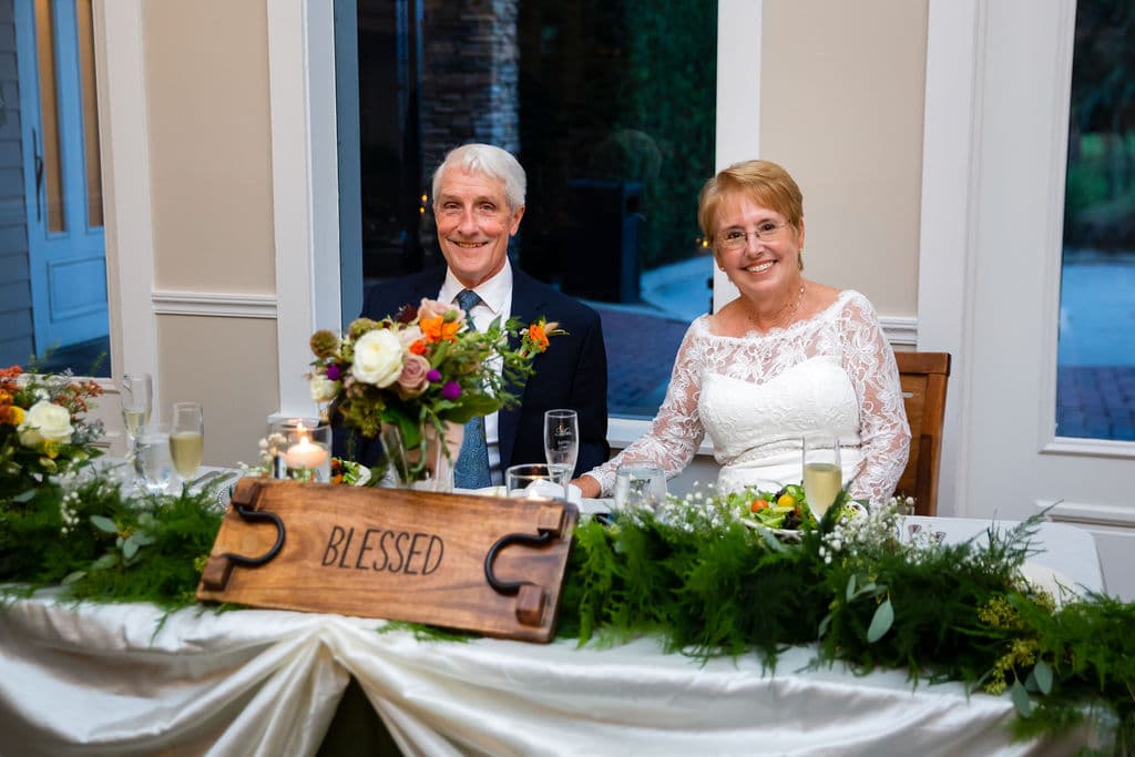 smiling bride and groom at sweetheart table with long green garland and wooden blessed sign in front and smaller floral arrangement behind the sign