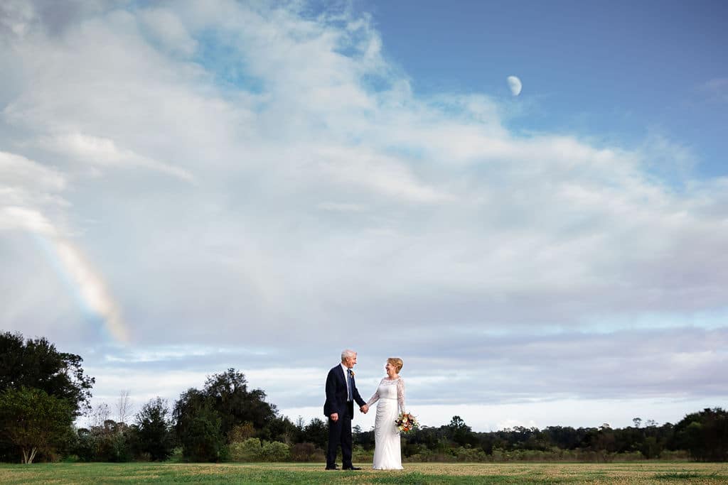 bride and groom in wedding pictures outside with fluffy white clouds and rainbow off to the left of the couple