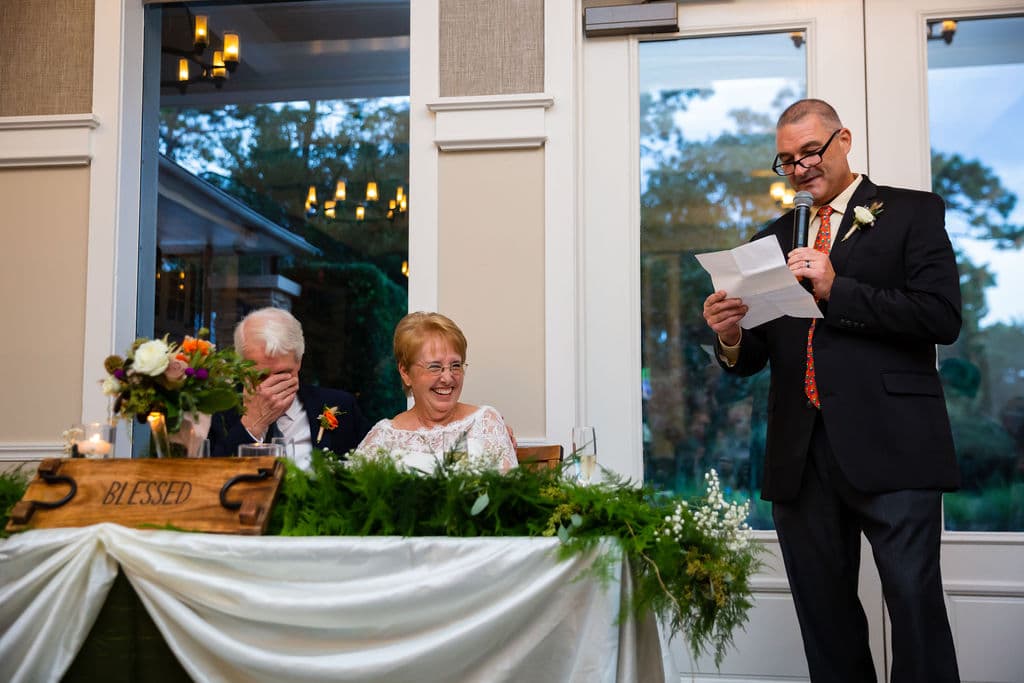 bride and groom sit at sweetheart table with garland and blessed wooden sign laughing while wedding guest holds microphone and reads from a piece of paper