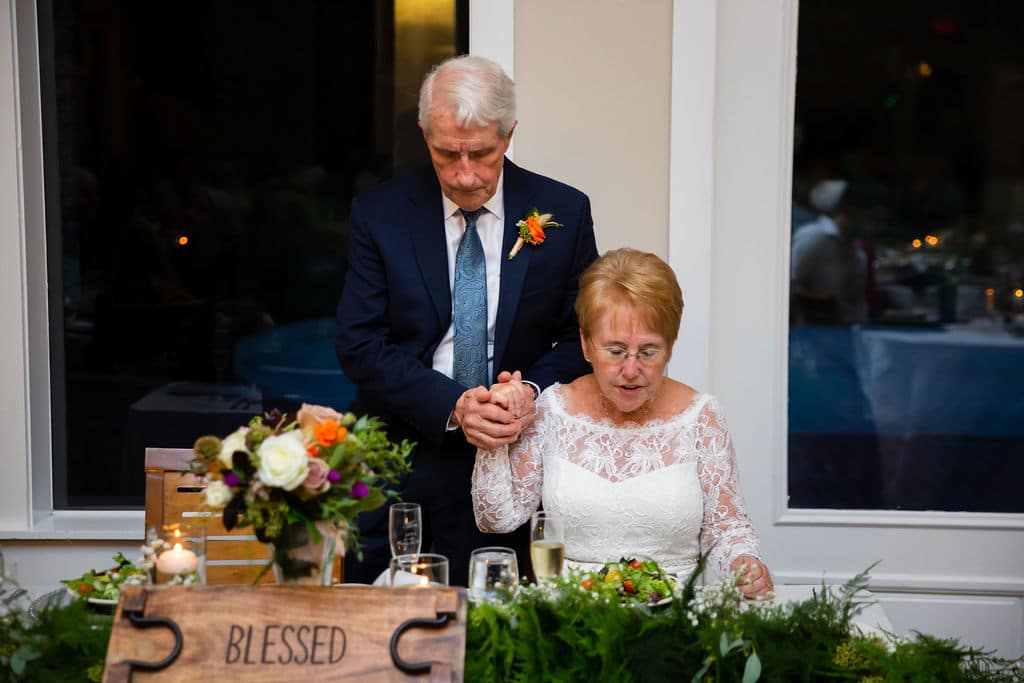 bride sits at table and groom stands behind holding hands over her right shoulder while bowing heads in prayer
