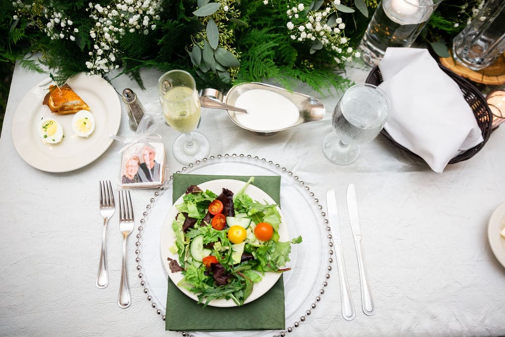 place setting at wedding reception with clear glass charger silver flatware vivid colored salad on top of charger with champagne glass and dressing boat