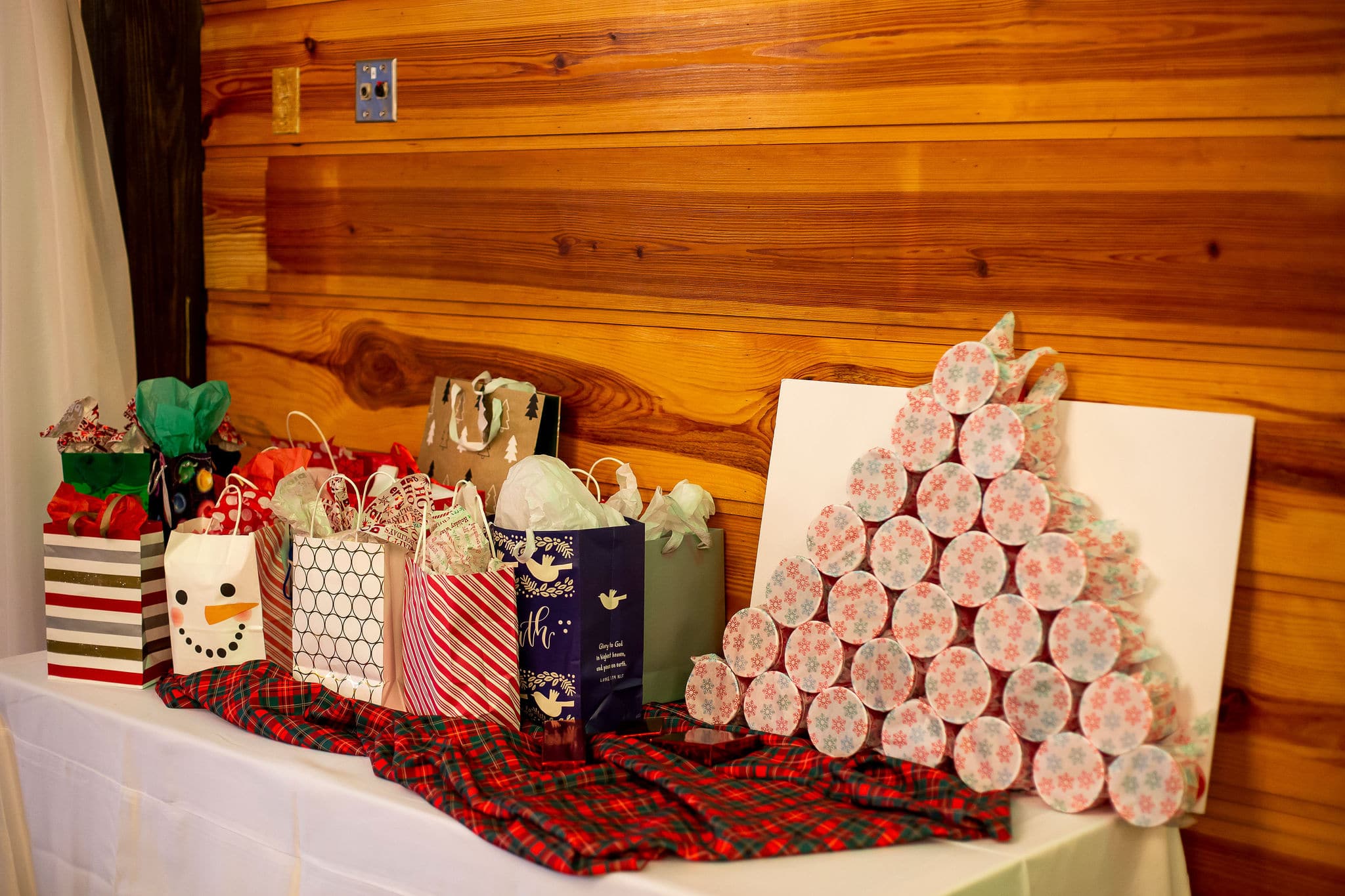 table in front of wooden wall with many christmas gift bags on top and a punch the hole game next to it on top of plaid linen