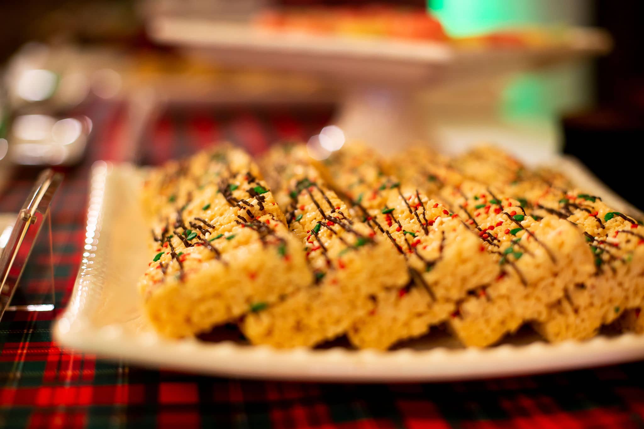 close up of rice krispie treats with chocolate drizzle and green white and red sprinkles on white plate sitting on top of plaid linen