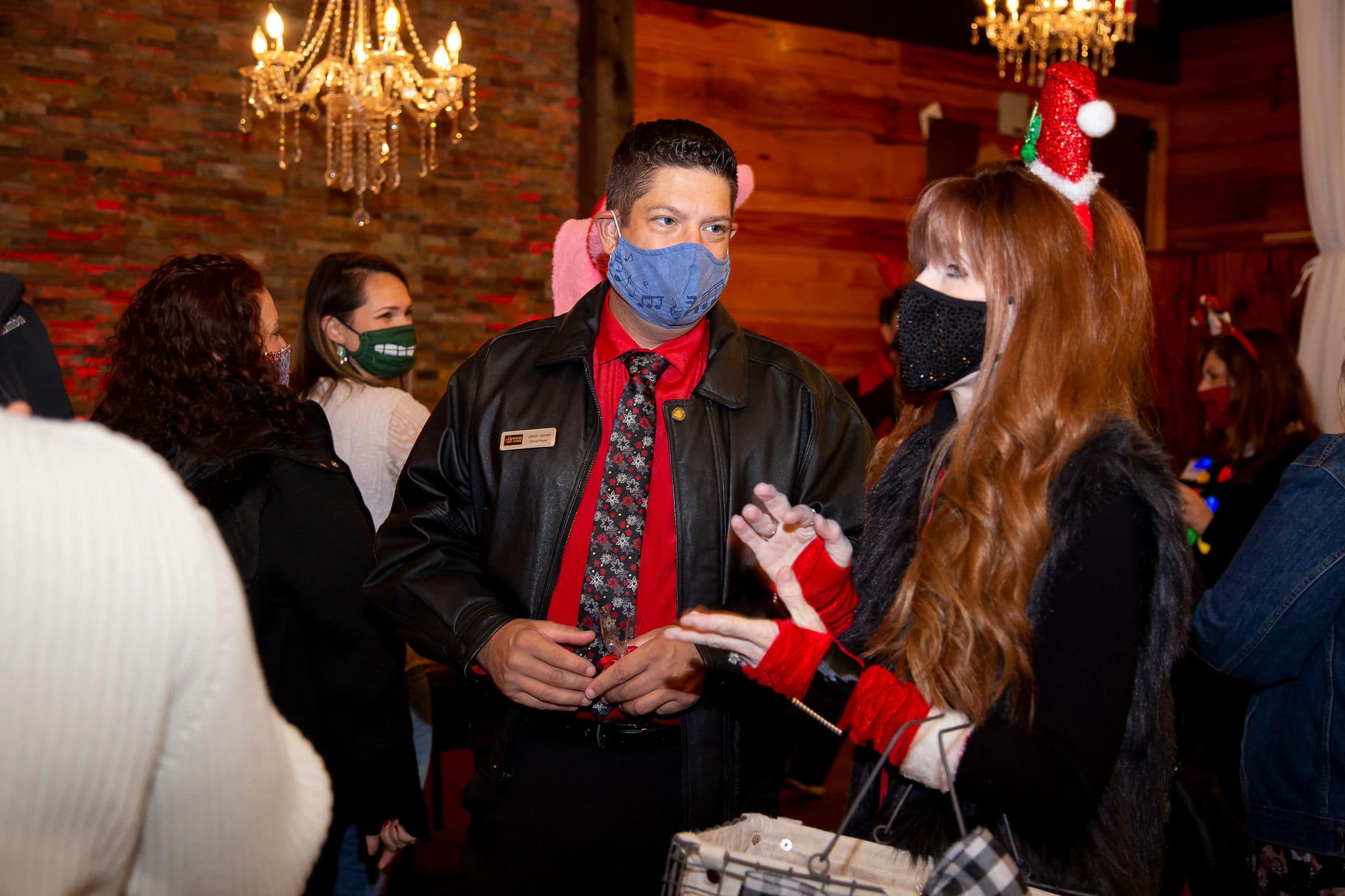 red haired woman wearing santa hat headband and santa-like gloves stands next to man wearing black jacket and red shirt with a holiday tie