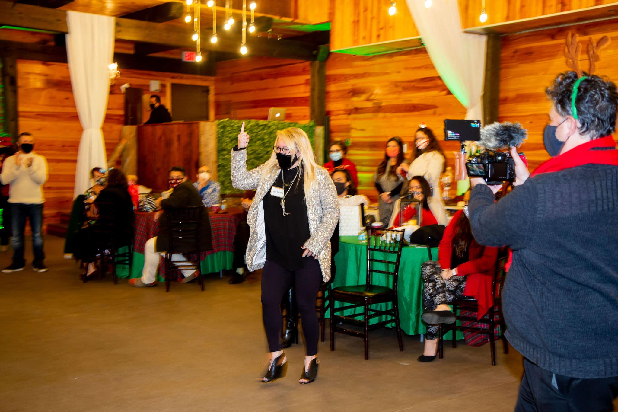blonde haired woman stands wearing all black pants top shoes and face covering with gold glitter jacket while other holiday party guest sit around her at table and videographer films the reaction