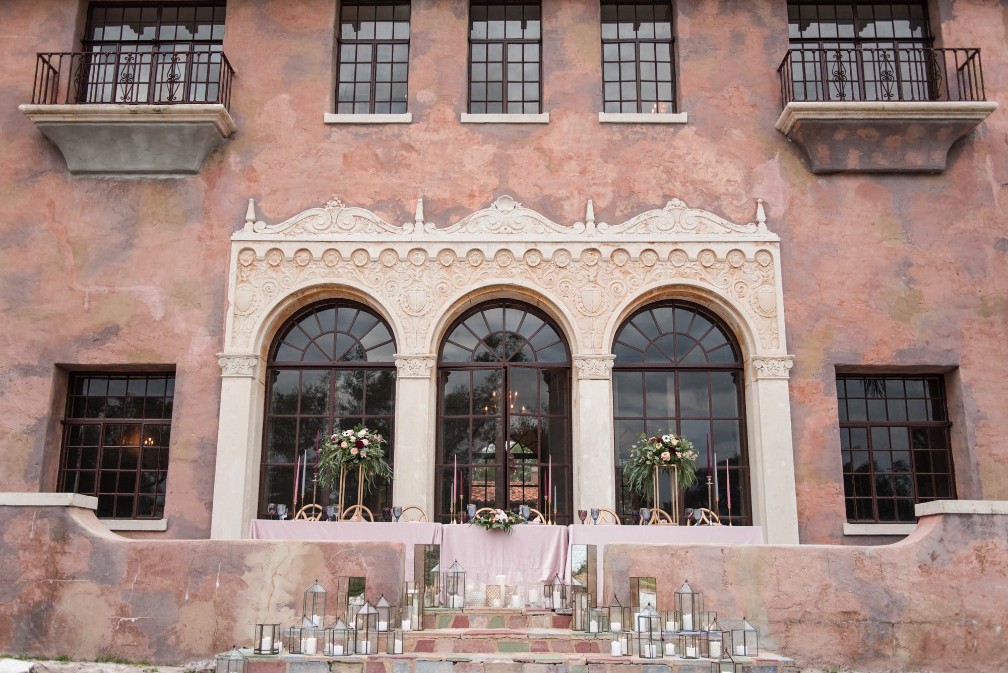 image of outside of historic building with wrought iron railings on balconies and three big windows in front of it decorated with table with light pink linen and candles on stairs going down