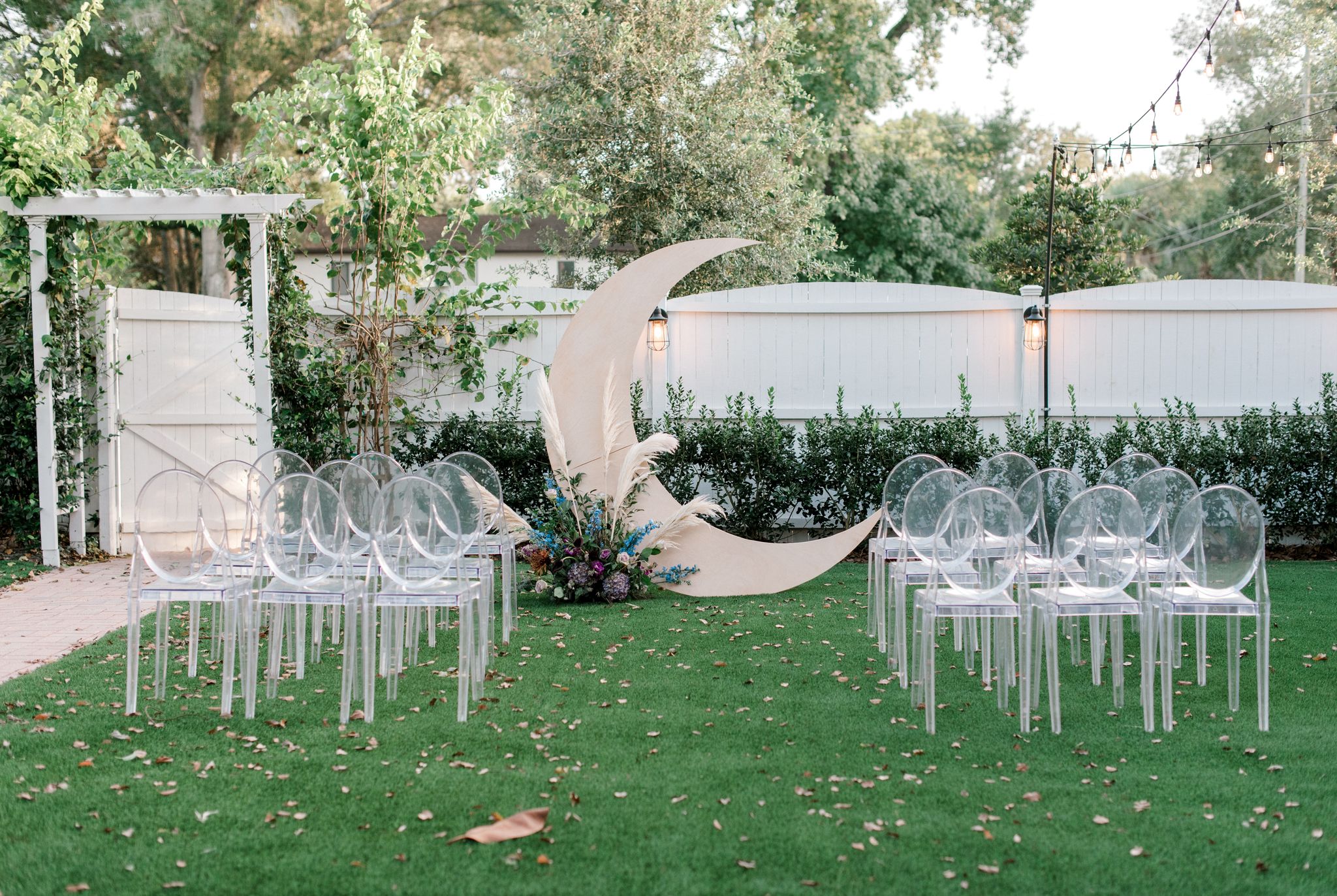 moon shaped arch on green lawn with clear chairs set up for wedding ceremony space in front of white fence