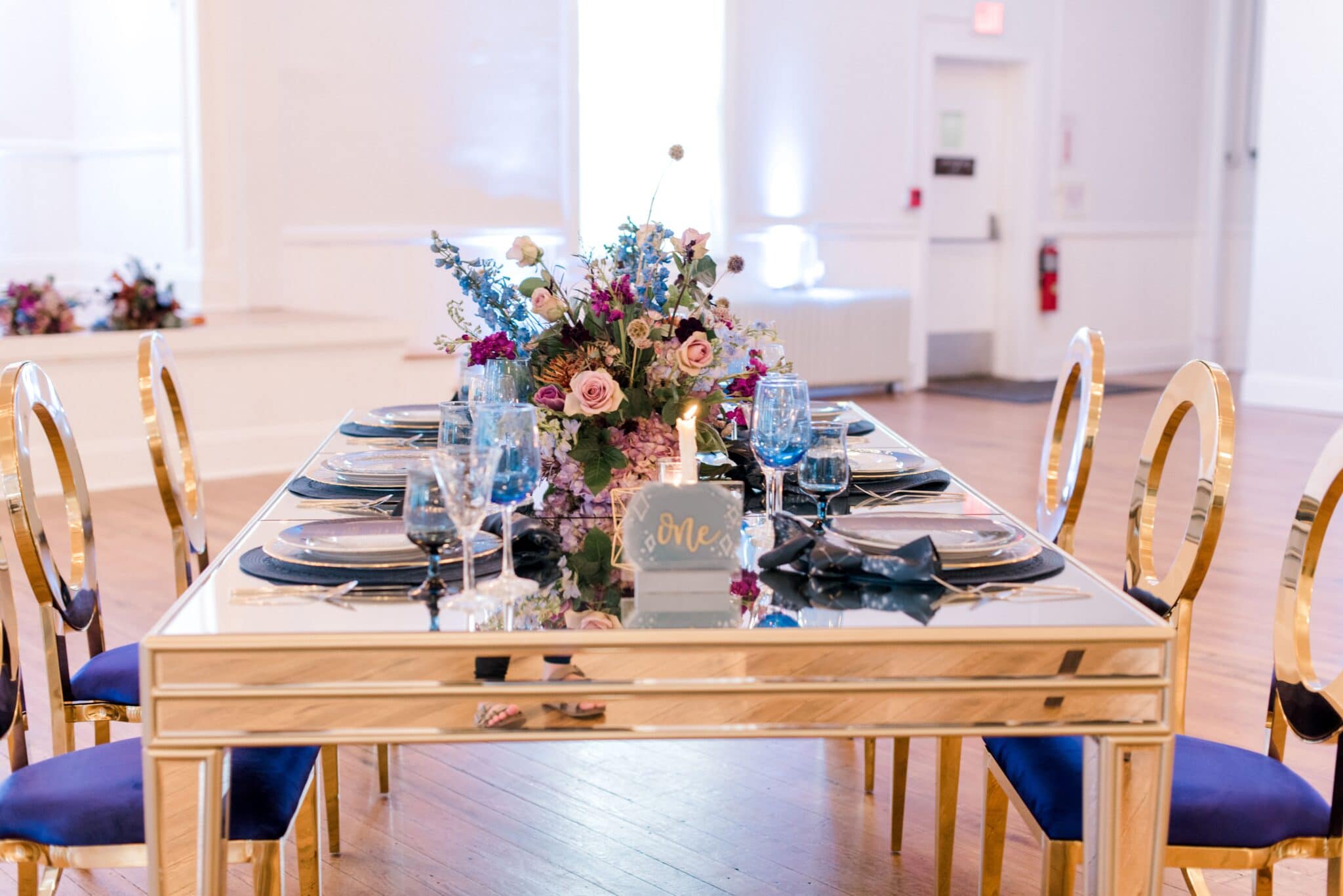 golden table with gold and blue padded chairs and the tabletop decorated with table number and tablewear along with floral arrangement