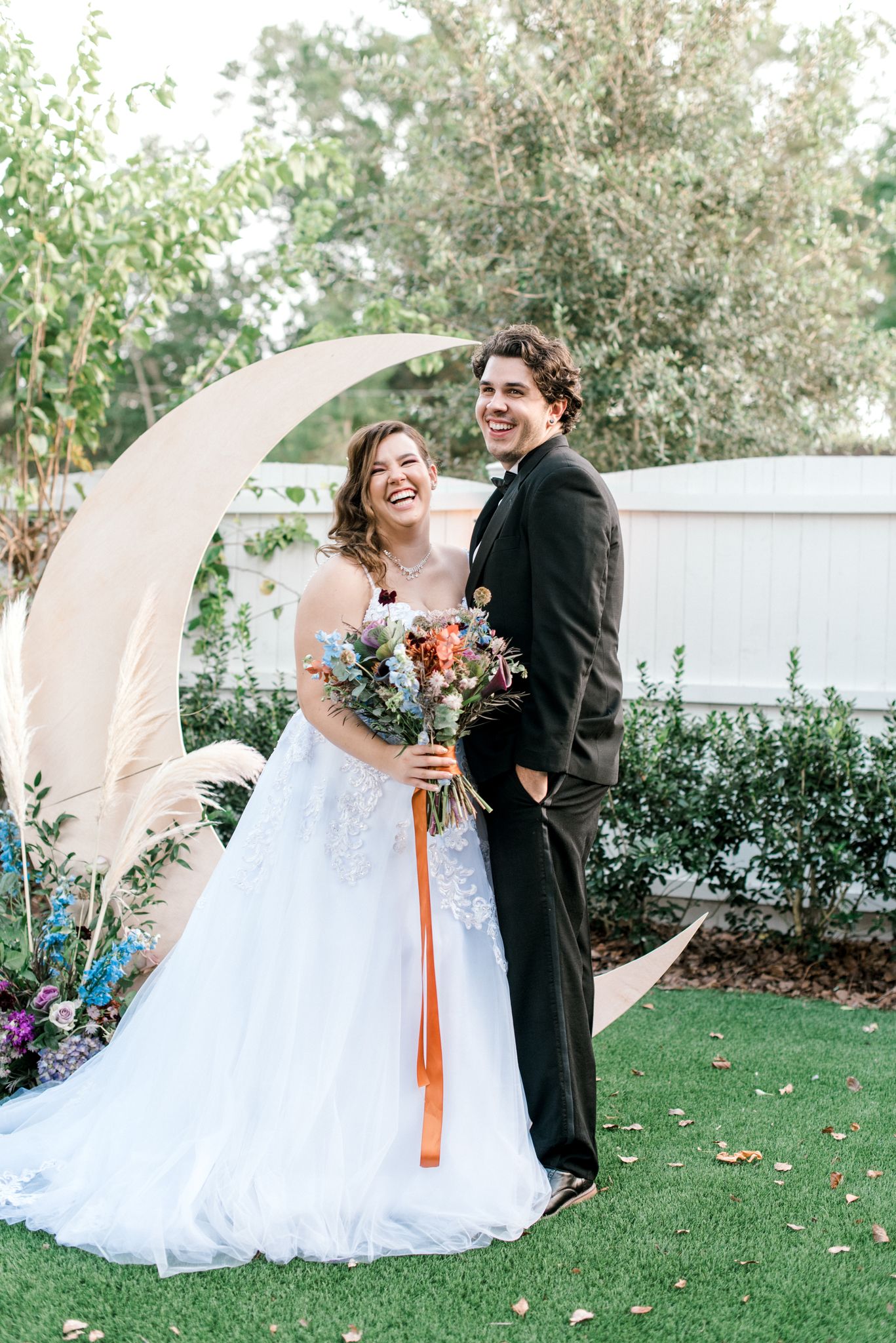 bridle holds bouquet with long orange ribbon next to groom in front of moon shaped ceremony arch