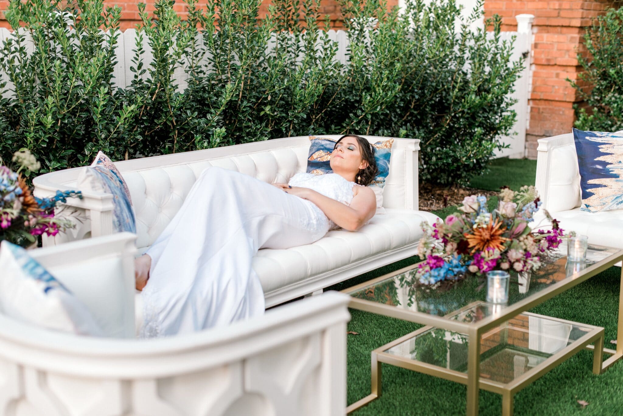 bride lays on cream colored couch in white wedding gown near a display of lounge furniture set up at wedding venue with flower arrangement on coffee table