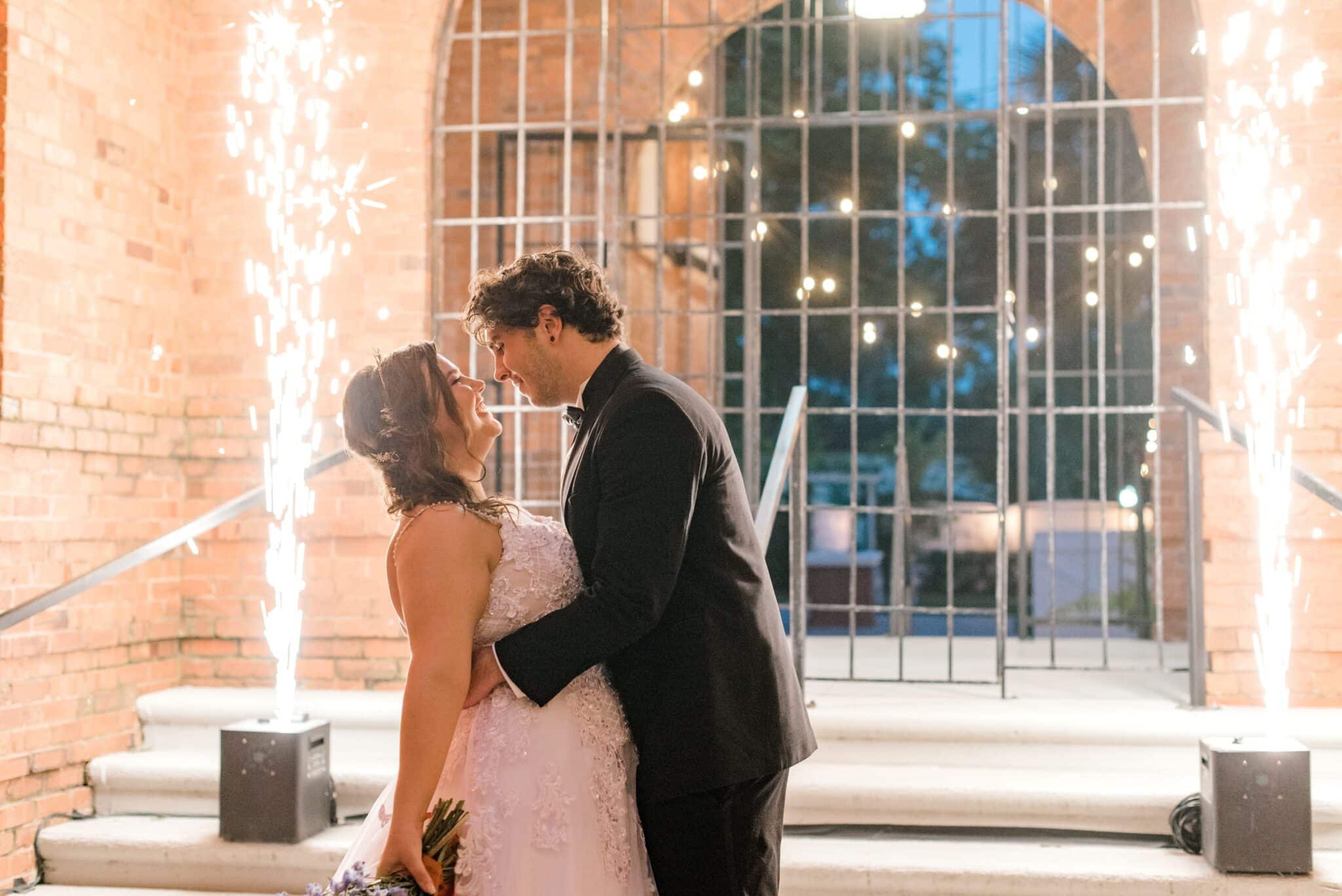 bride and groom in wedding portrait in front of wrought iron gate and two sparkler machines at nighttime