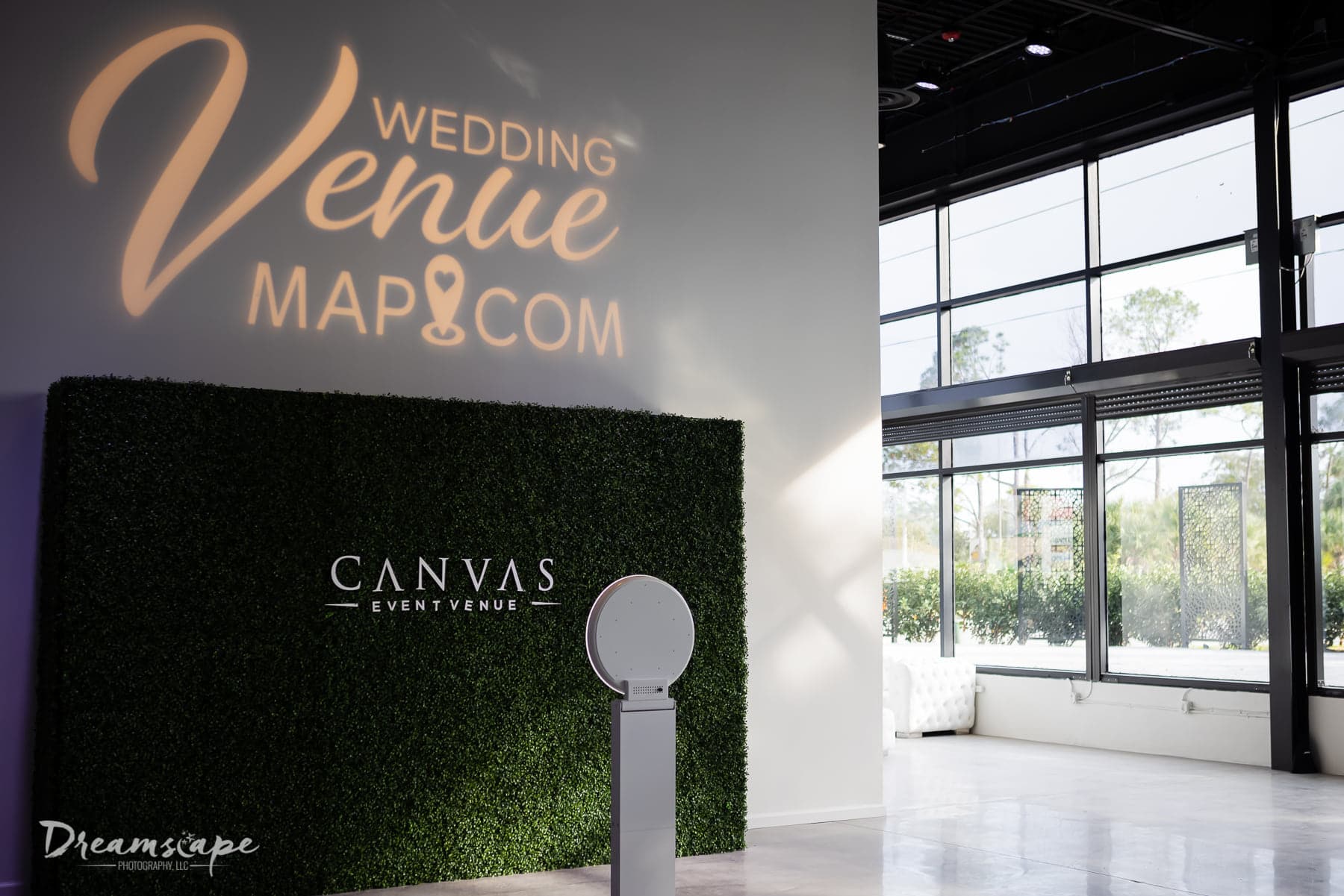 empty photo booth area with hedge wall with logo of venue on the wall and a different logo projected on the wall above the greenery with white photo booth stand in front