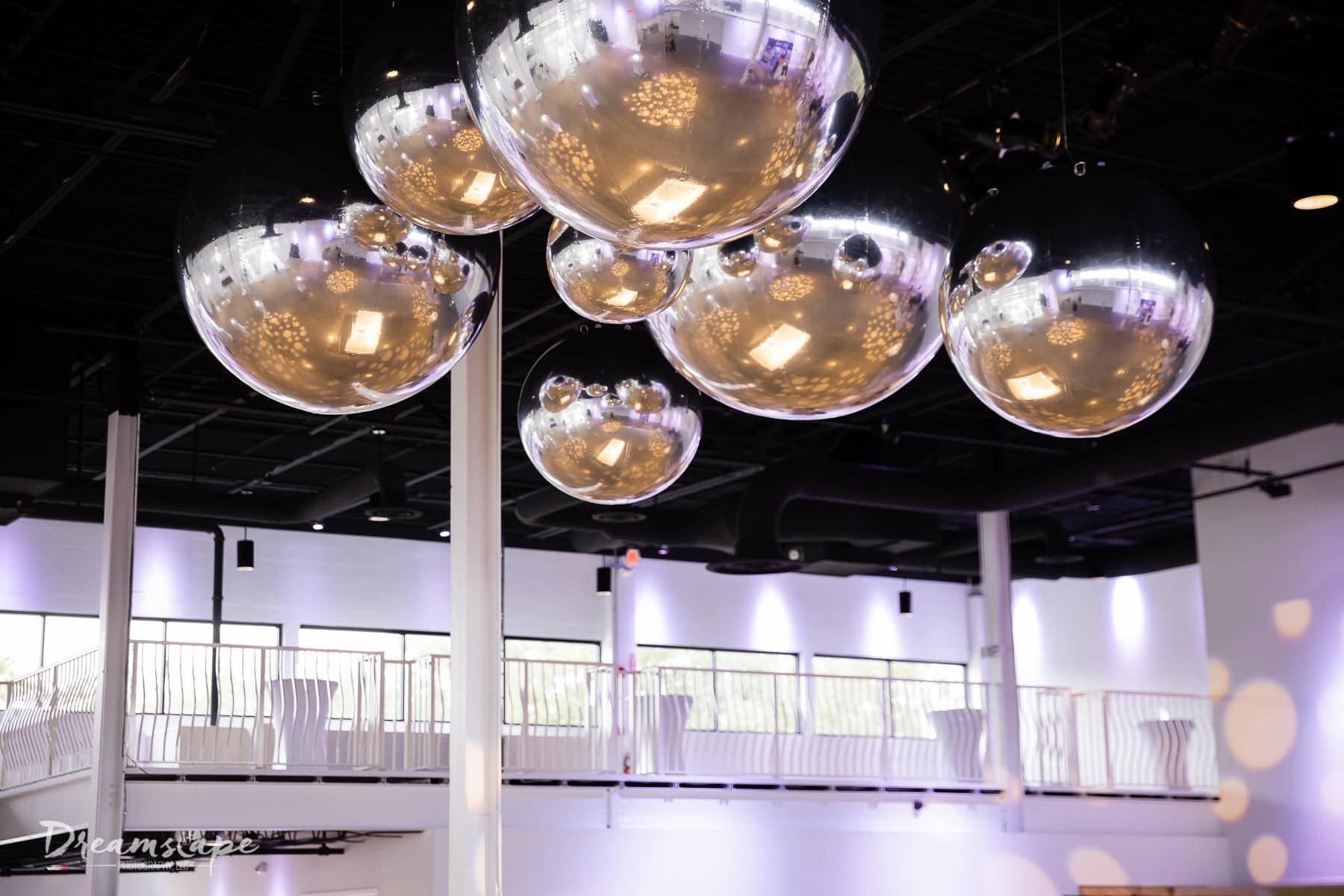 looking at balcony area of venue with white railing and tall cocktail tables set up on the balcony with big silver balls hanging from the ceiling