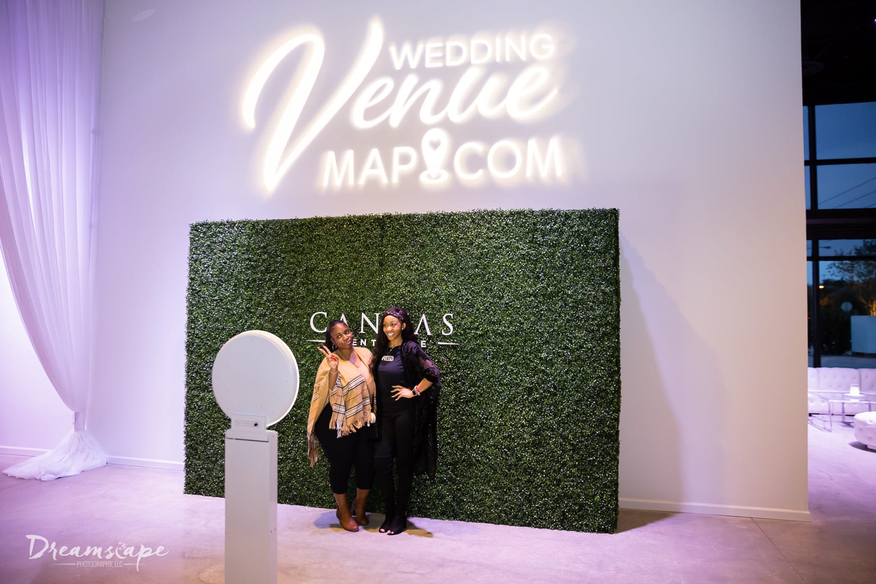 two woman stand with arms around each other in front of green hedge wall with logo projected above it for photo booth picture