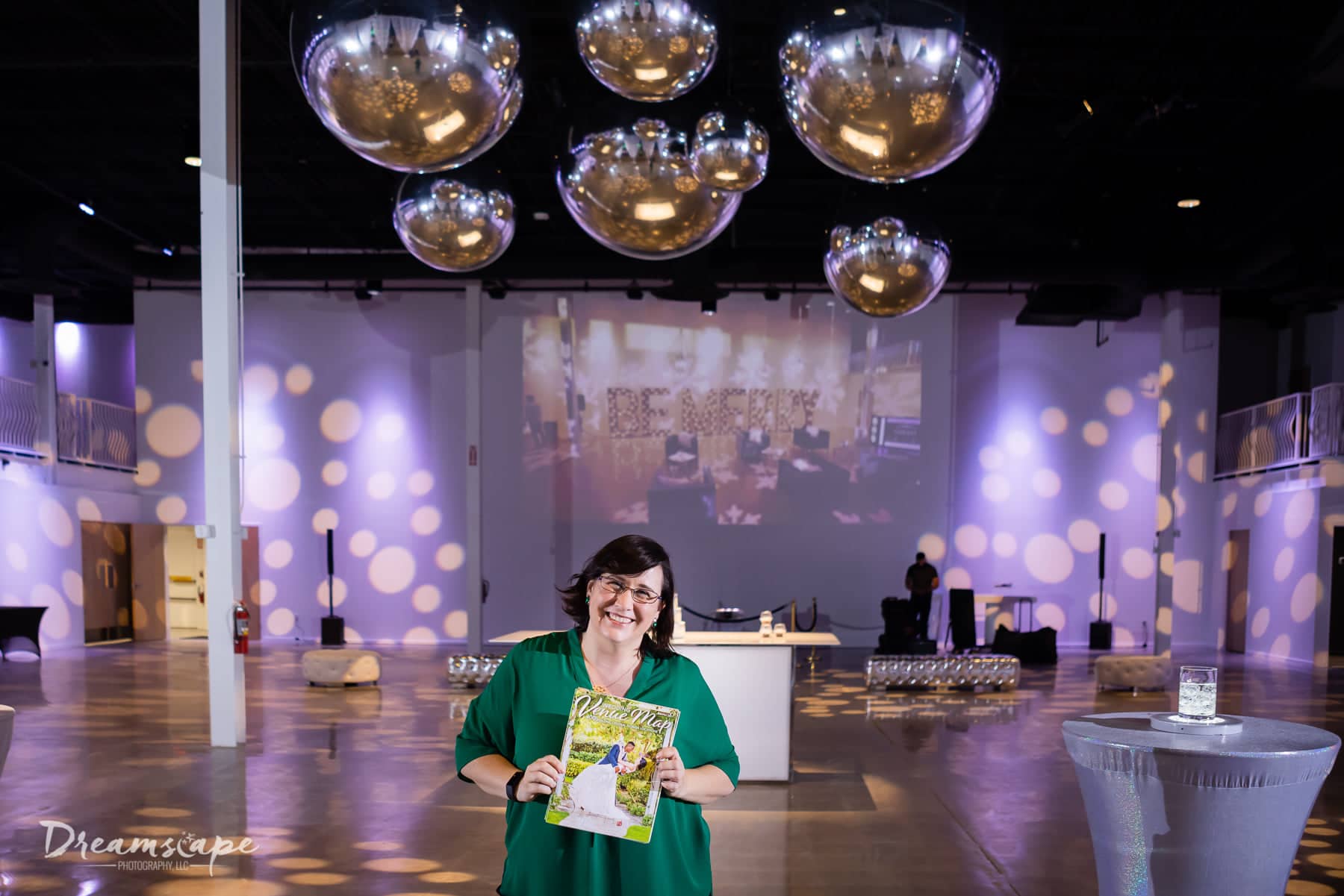 woman with glasses and short brown hair wearing green shirt holding map in front of venue space with big silver balls hanging from ceiling new map celebration at Canvas Event Venue