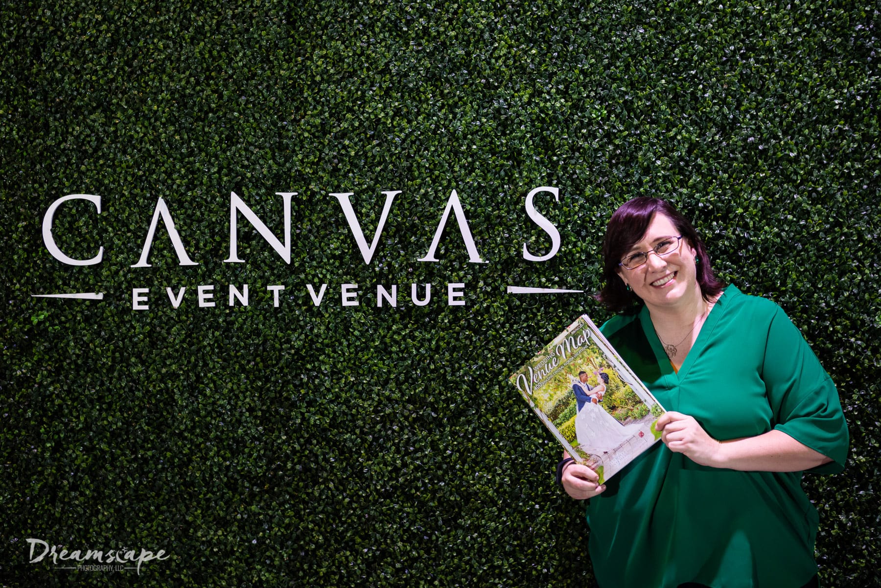 woman in green blouse wearing glasses and holding a printed and folded map smiling in front of hedge wall with canvas event venue logo on it