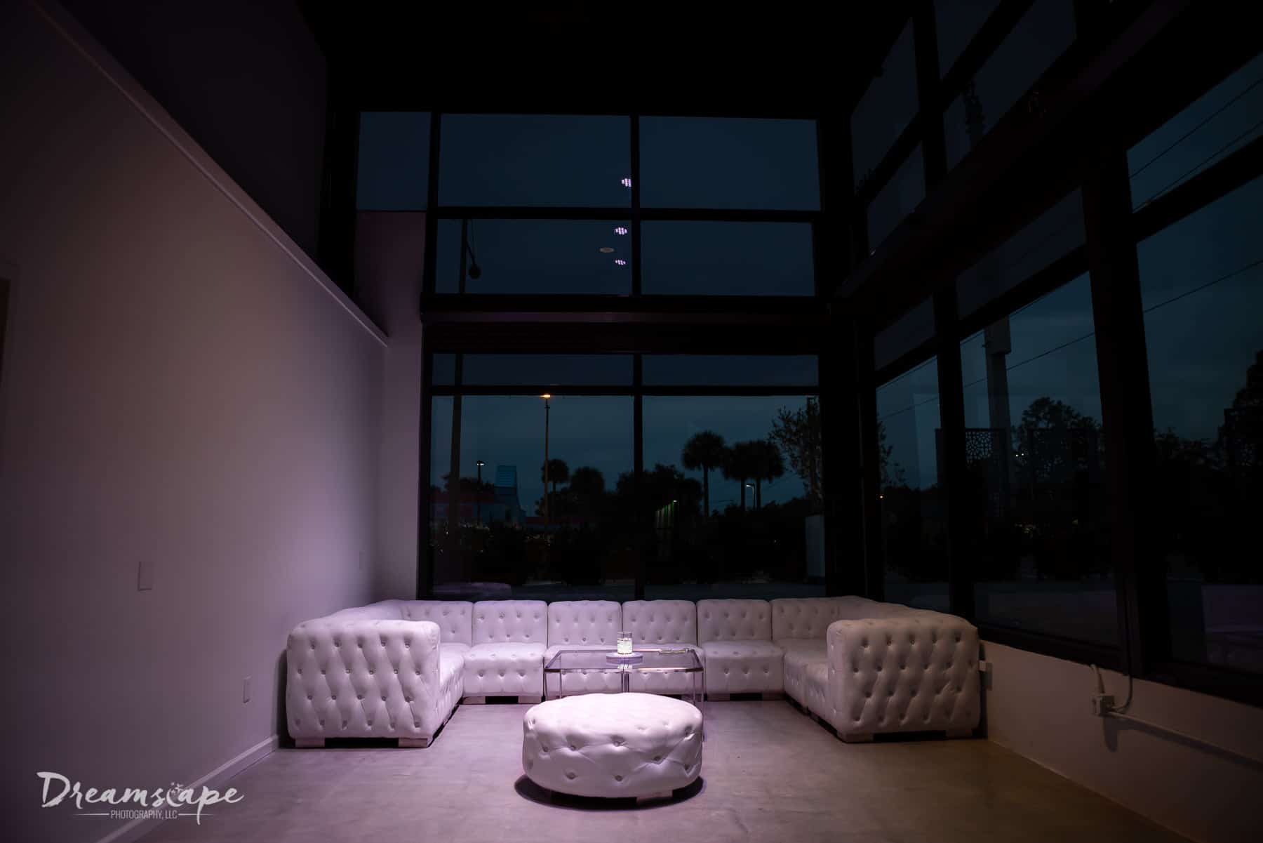 nighttime image of white leather sofas with clear coffee table in front of large windows and white wall to the left
