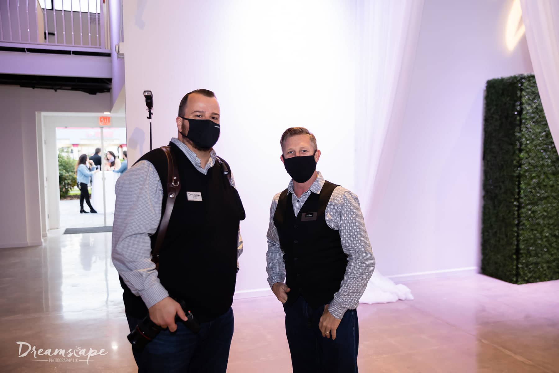 two men wearing the same long sleeve and sweater vest combination stand next to each other with black face masks on with open door to outside behind them
