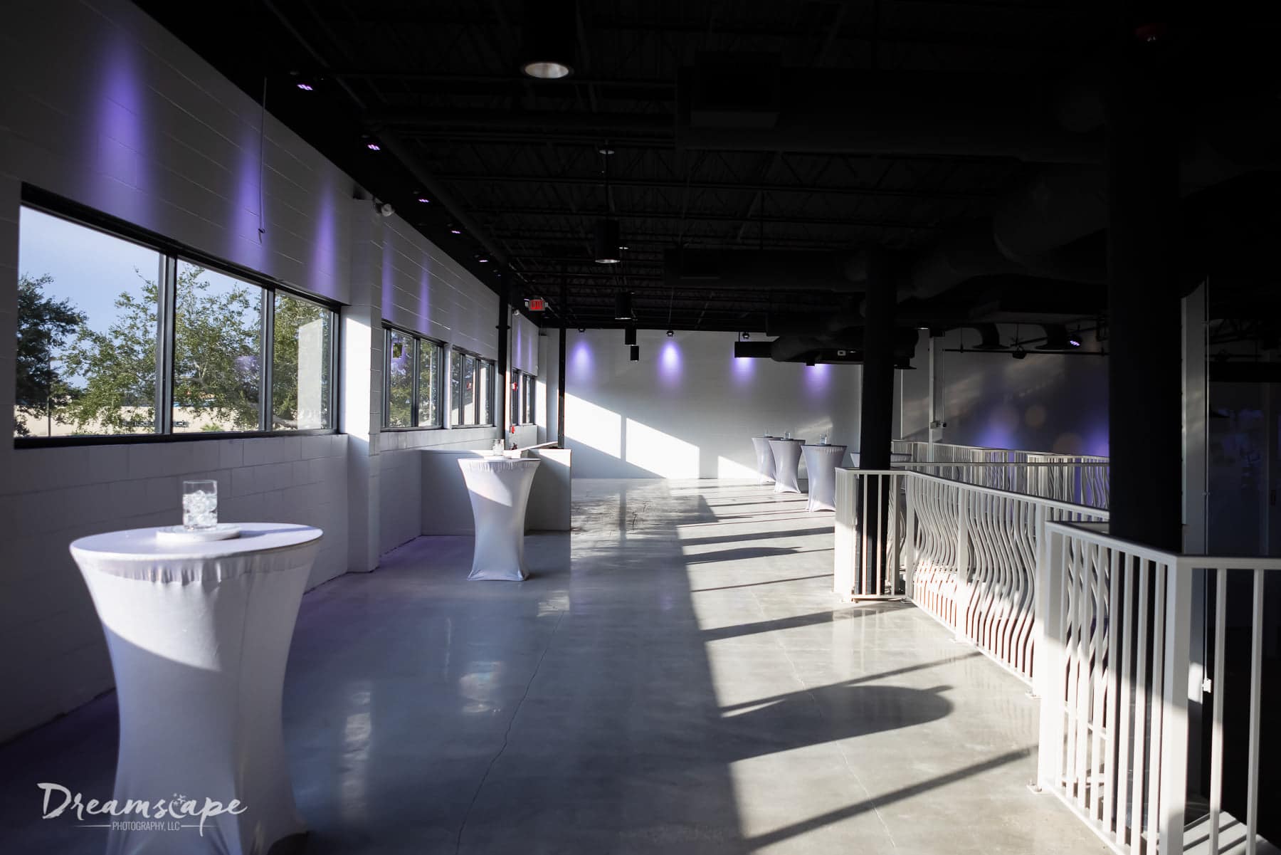 balcony of venue with windows on the left side of the space and to the right the white railing overlooking the main event space and cocktail tables set up throughout the space