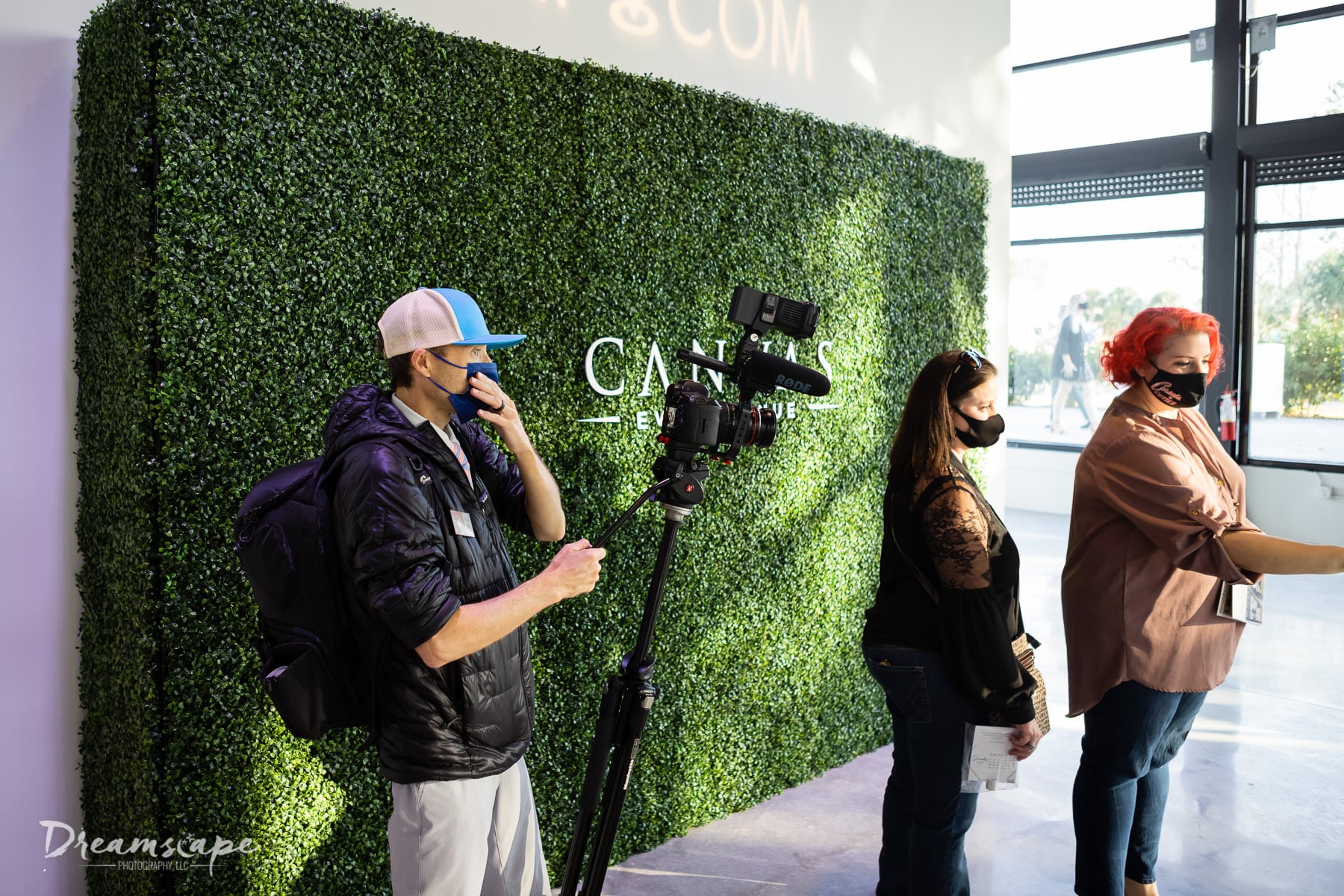 man wearing hat and big black jacket stands next to hedge wall and two women using photo booth while holding videographer equipment