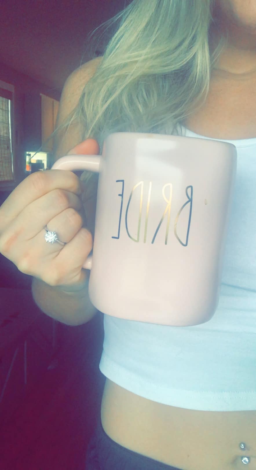 woman holds dusty pink colored mug with the word bride written on it in gold letters with left hand showing engagement ring wearing a white shirt