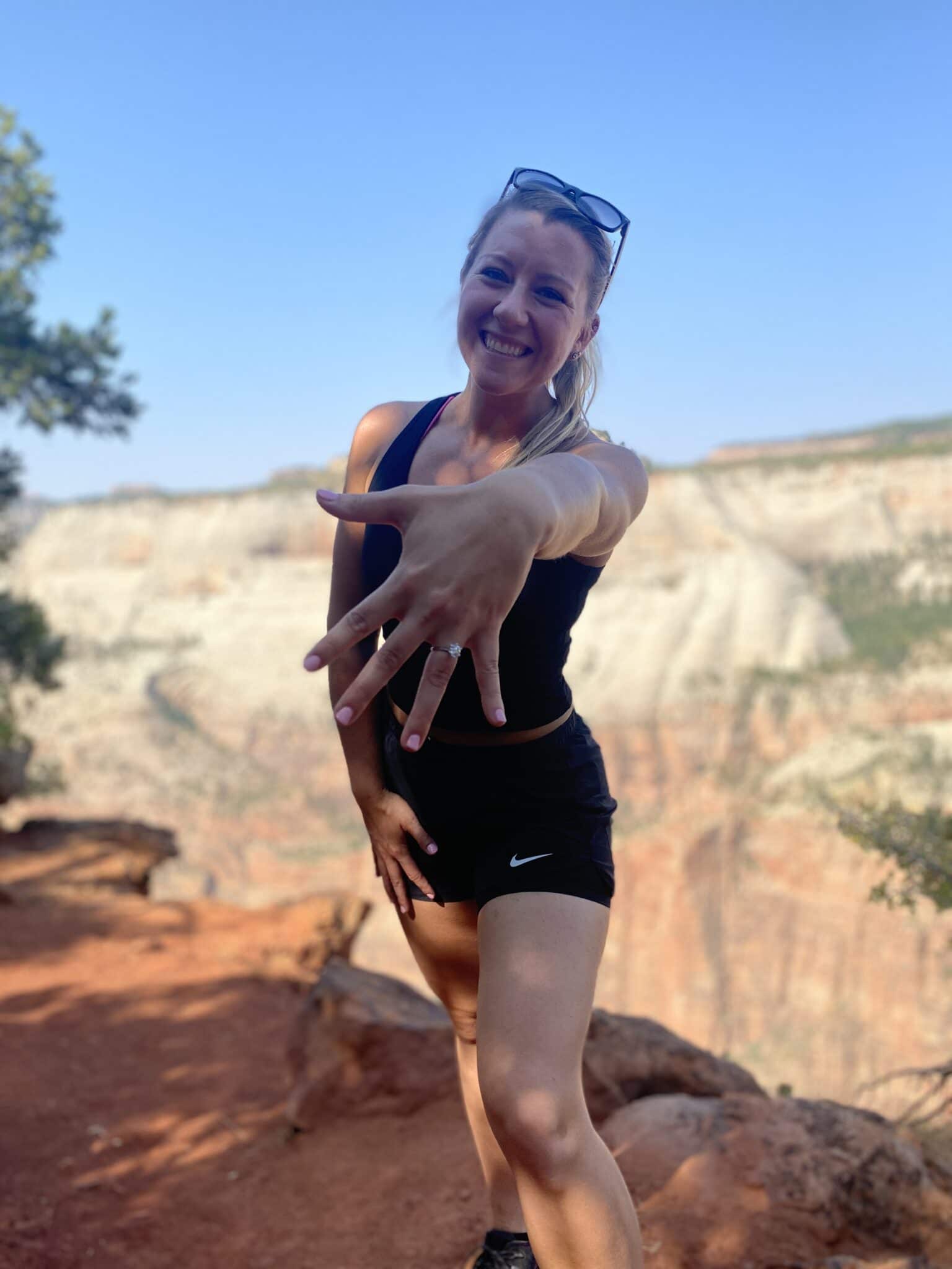girl wearing black shorts and tank top stands at the top of mountain near cliff holding out her ring finger towards camera to show off engagement ring