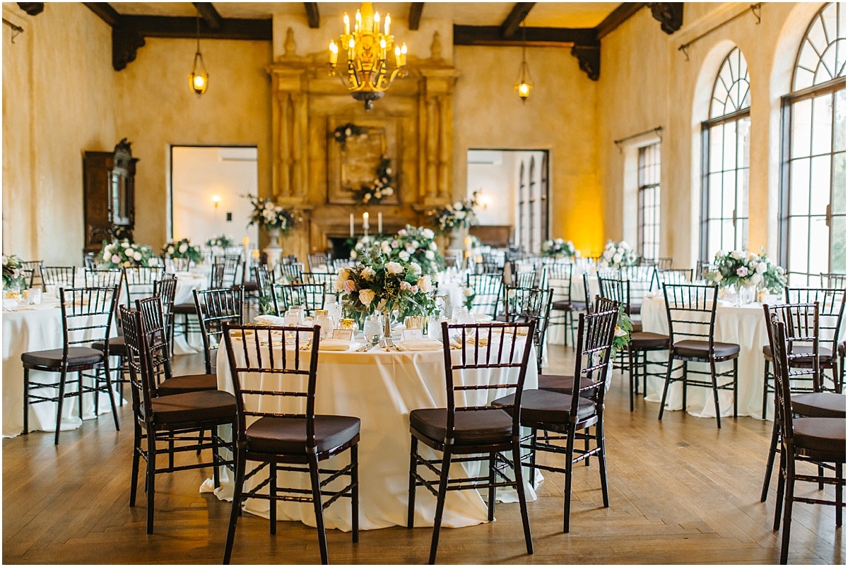 indoor ballroom set up with round table and white linen with mahogany chiavari chairs and short floral arrangements in the center of each table