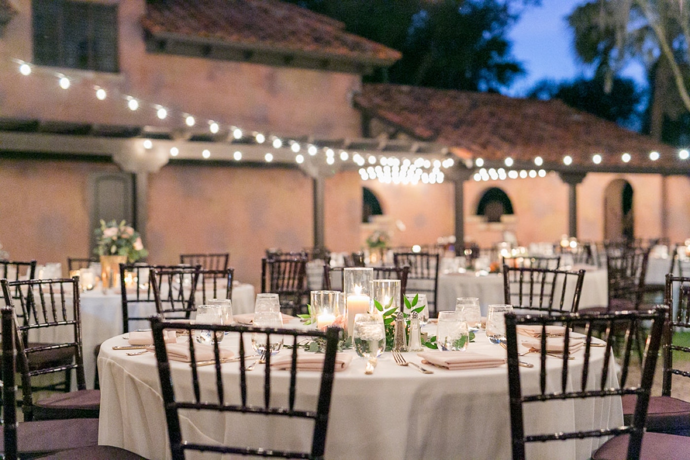 close up of tables and chairs set at night for wedding reception in outdoor space with market lights overhead and white linen on the tables and candles with some greenery