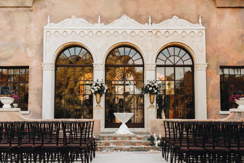 ceremony space set up with tall stands and florals in gold vases at the alter with a tiny table with white linen and mahogany chiavari chairs the venue with three huge windows