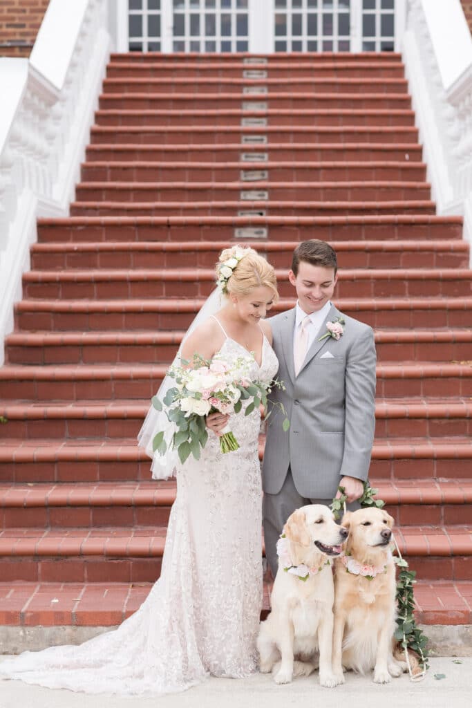 bride and groom standing at bottom of outdoors steps with groom holding leashes of their two dogs