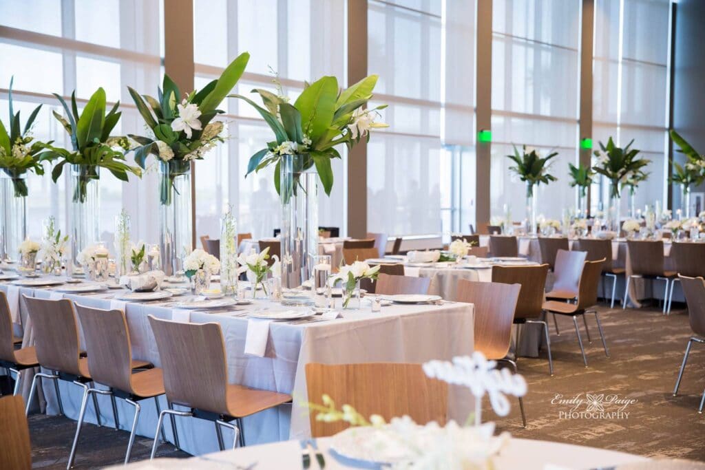 long tables set up with white place settings and tall green tropical floral arrangements in ballroom with large windows