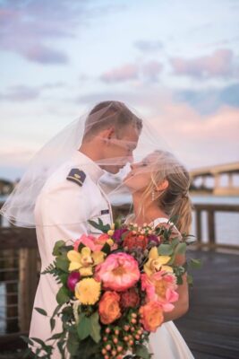 bride and groom both under bride's veil and holding large tropical bouquet by the water at sunset