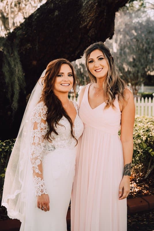 bride and maid of honor in their dresses with hair and makeup done standing outside under large tree