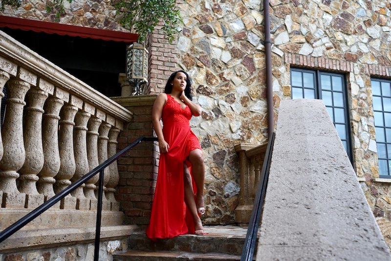 woman in red dress leaning on railing outside of old stone building with hair and makeup done by House of Sheen Glam Team