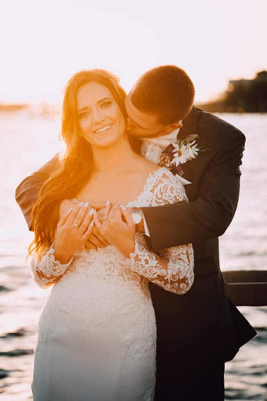 groom kissing his bride on their wedding day standing on a lake shore at sunset with brides make-up by House of Sheen Glam Team
