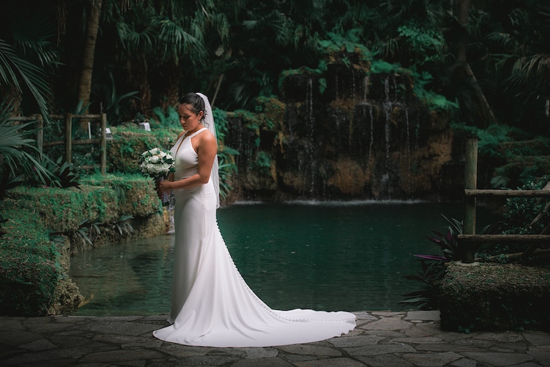 bride standing in front of pond surrounded by trees with a waterfall
