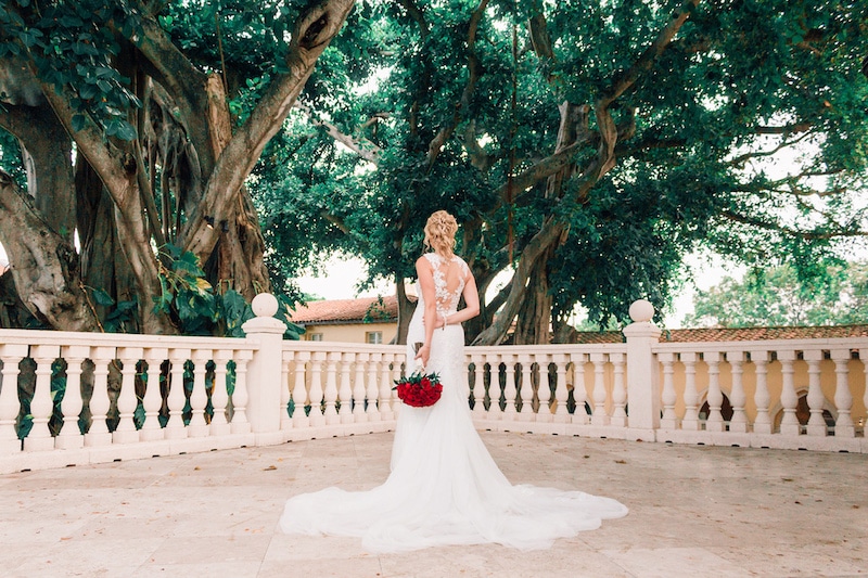 bride standing on outdoor patio holding red flower bouquet under cover of large old trees photo by Cona Studios