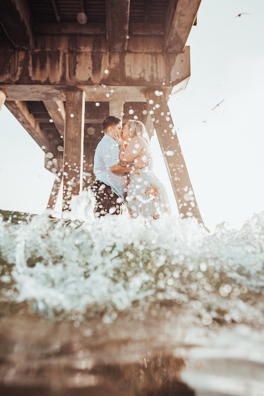 couple kissing while standing in the water beneath a pier photo by Cona Studios