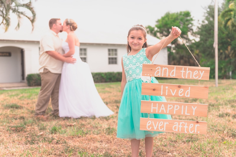 bride and groom kiss while young girl holds sign that reads 