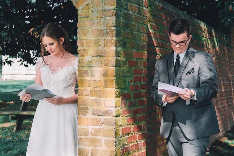 bride and groom standing on either side of a brick fence reading a note they read for each other photo by Cona Studios