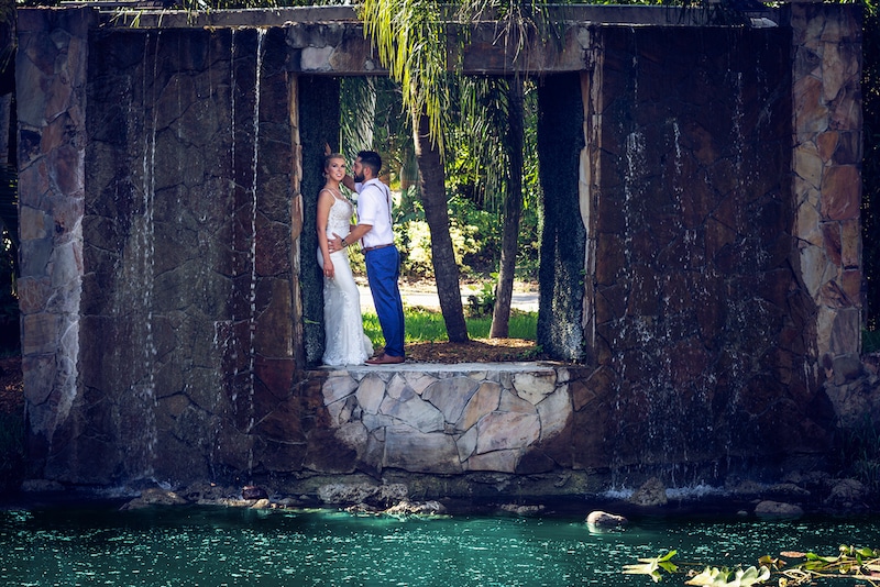 groom leaning in to kiss bride standing in stone cutout above a pond with waterfall photo by Cona Studios