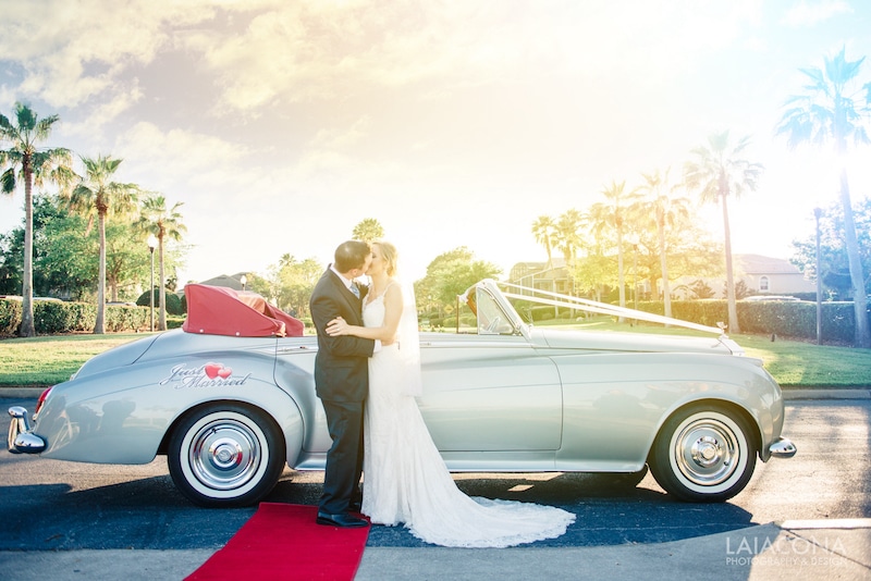 bride and groom kissing while standing in front of classic convertible with red carpet photo by Cona Studios