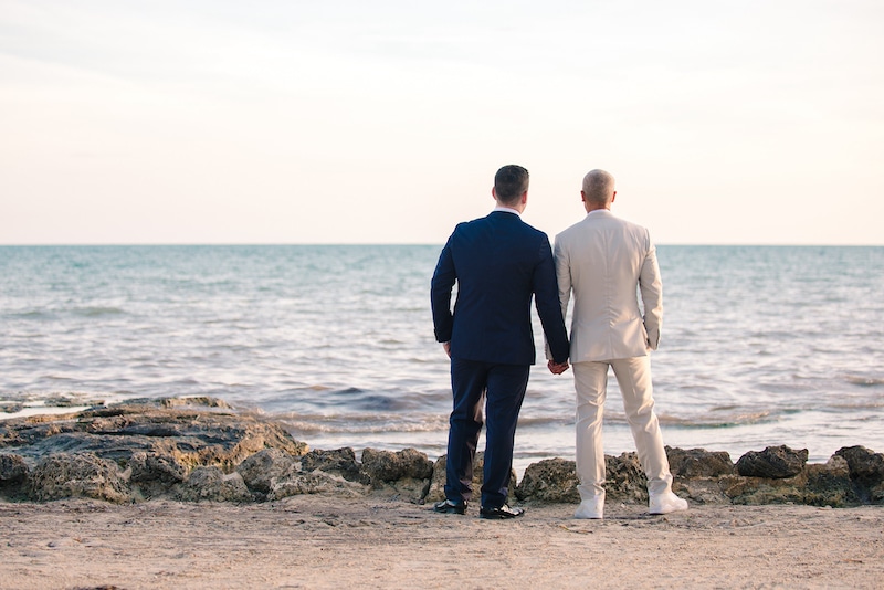 grooms holding hands standing on the beach overlooking the ocean photo by Cona Studios