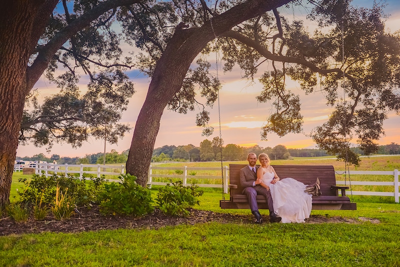 bride and groom sitting on long bench swing tied to tree on a farm with sun setting in background
