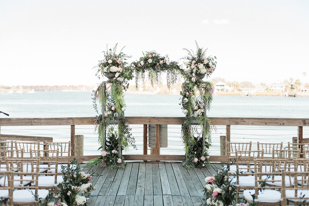 outdoor wedding ceremony set up on dock with white chairs and floral arch overlooking the water