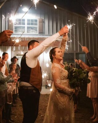 bride and groom holding hands and dancing in front of barn while guests hold sparklers around them after wedding