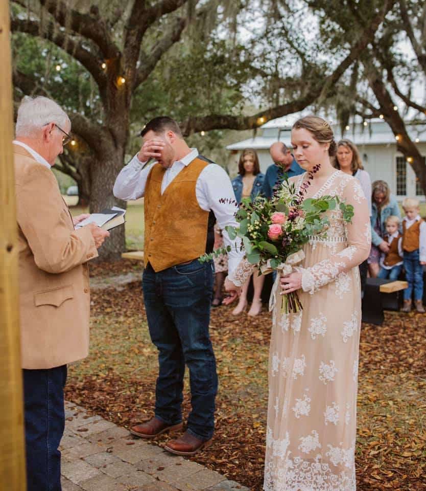 bride and groom praying with eyes closed during outdoor wedding ceremony under large oak trees on farm