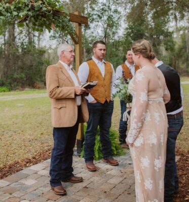 bride in white lace dress and groom with black vest standing at outdoor wedding ceremony with officiant under wooden archway