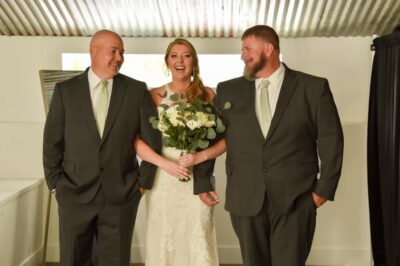 bride holding white bouquet and smiling while being escorted by two men in gray suits