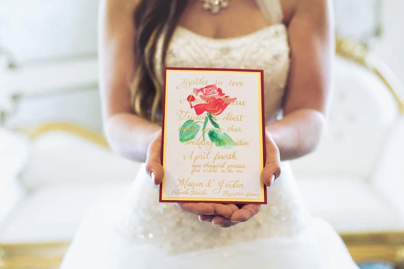 bride holding custom invitation featuring a rose, calligraphy, and red and gold colors